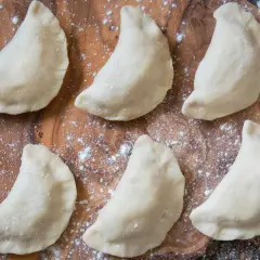 Sześć surowych pierogów z jabłkami ułożonych na drewnianej desce, oprószonych mąką.
