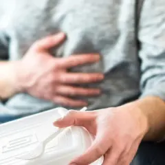 Mężczyzna trzymający plastikowe pudełko z jedzeniem i trzymający się za brzuch.