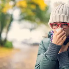 Kobieta w czapce z pomponami i okularach chusteczką wyciera nos na tle jesiennego parku.