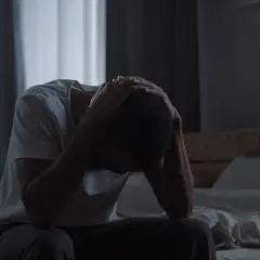 Mężczyzna siedzi na łóżku, trzymając się za głowę, w przyćmionym świetle.