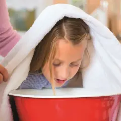 Dziewczynka pochyla się nad miską z gorącą wodą, przykryta ręcznikiem, podczas inhalacji.