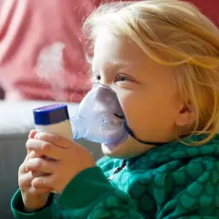 Dziecko z blond włosami używa inhalatora, z którego wydobywa się para.