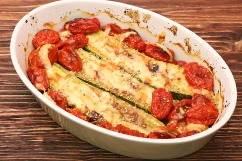 Zapiekana cukinia z pomidorami i serem, posypana sezamem, w białej naczyniu żaroodpornym na drewnianym tle.
