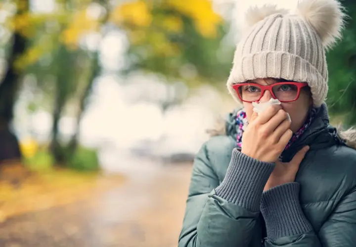Kobieta w czapce z pomponami i okularach chusteczką wyciera nos na tle jesiennego parku.