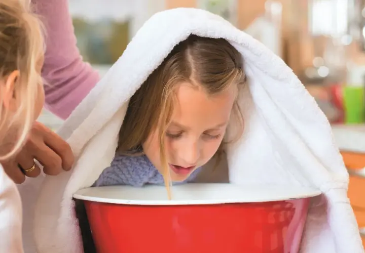 Dziewczynka pochyla się nad miską z gorącą wodą, przykryta ręcznikiem, podczas inhalacji.