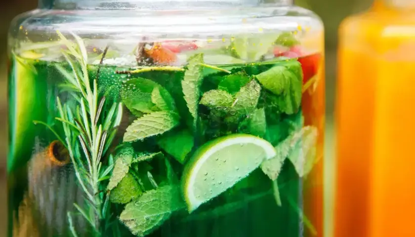 Orzeźwiający sok z mięty w szklanym słoju, z dodatkiem limonki i rozmarynu. Obok pomarańczowy napój.