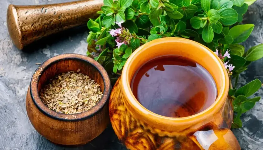 Napar z majeranku w ceramicznym kubku, obok drewnianej miseczki z suszonym majerankiem i świeżych gałązek z kwiatami.