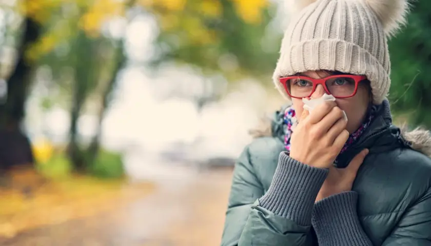 Kobieta w czapce z pomponami i okularach chusteczką wyciera nos na tle jesiennego parku.