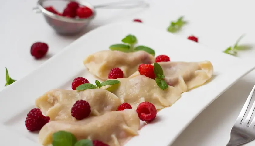 Pyszne pierogi z malinami, udekorowane świeżymi malinami i listkami mięty, prezentują się apetycznie na białym talerzu.