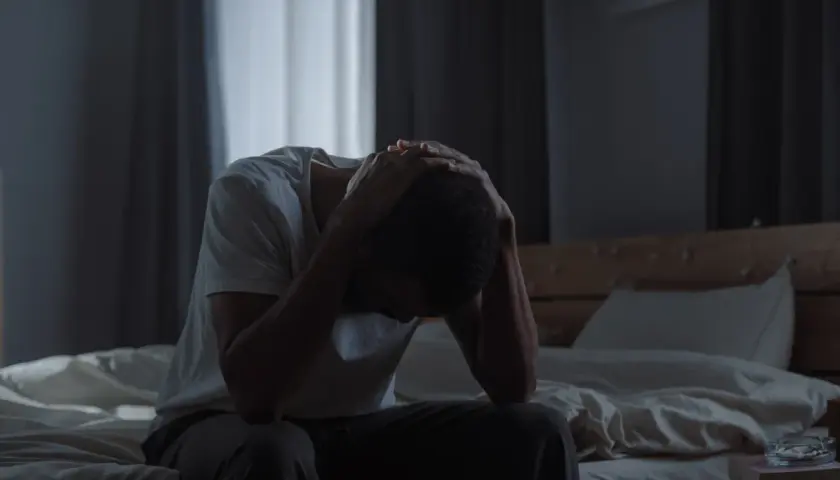 Mężczyzna siedzi na łóżku, trzymając się za głowę, w przyćmionym świetle.