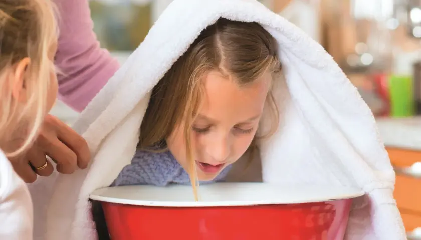 Dziewczynka pochyla się nad miską z gorącą wodą, przykryta ręcznikiem, podczas inhalacji.