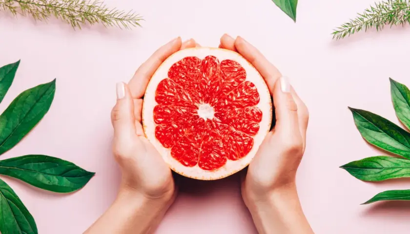 Dłonie trzymają połówkę grejpfruta, otoczone zielonymi liśćmi. Naturalne piękno i świeżość, jak wyciąg z pestek grejpfruta.