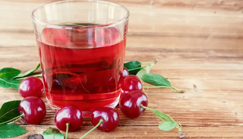 Szklanka z rubinowym kompotem z wiśni na zimę, otoczona świeżymi owocami i listkami na drewnianym tle.