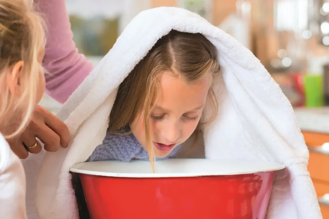 Dziewczynka pochyla się nad miską z gorącą wodą, przykryta ręcznikiem, podczas inhalacji.