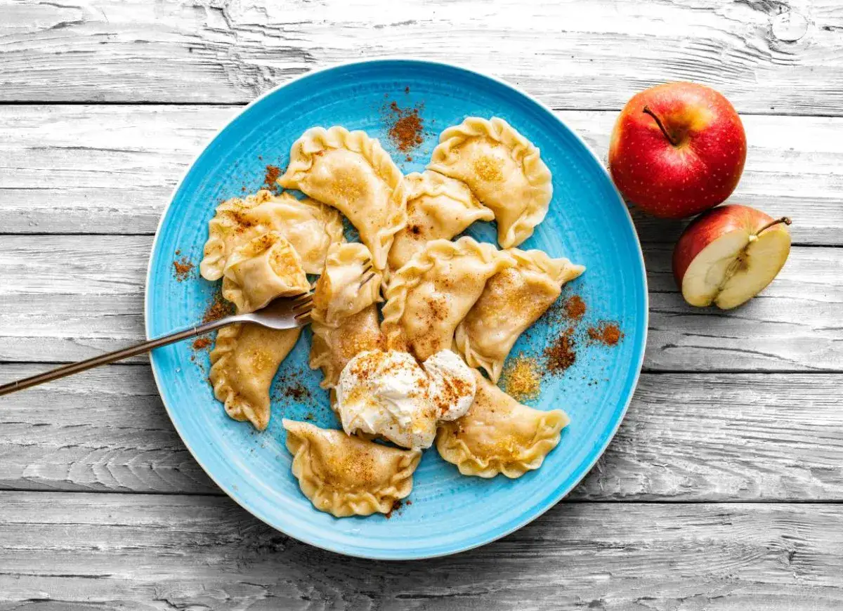 Pyszne pierogi z jabłkami, posypane cynamonem, z porcją śmietany. Obok leżą świeże jabłka.